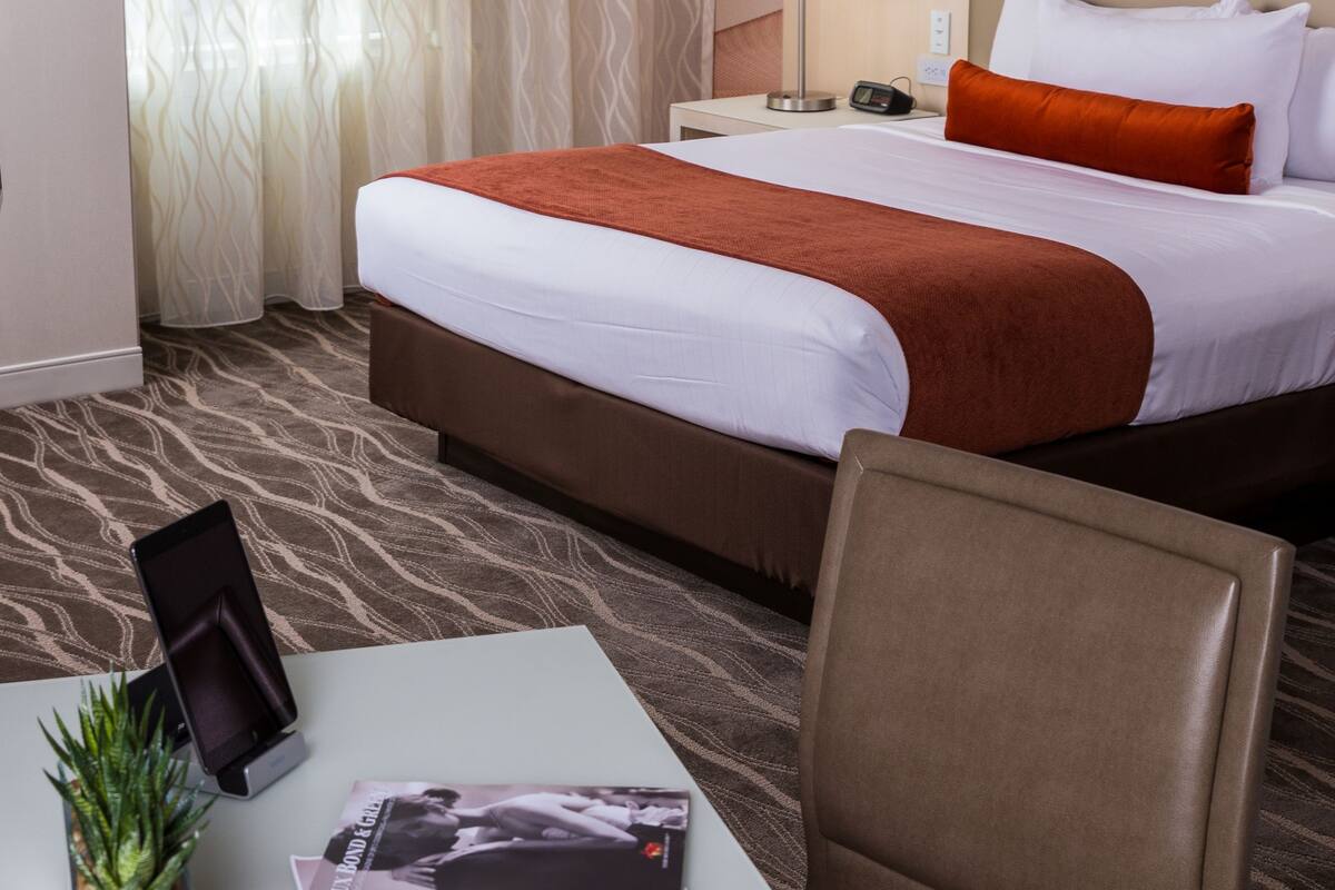 Quarto de hotel com cama, escrivaninha com luminária, cadeira e uma pequena planta.