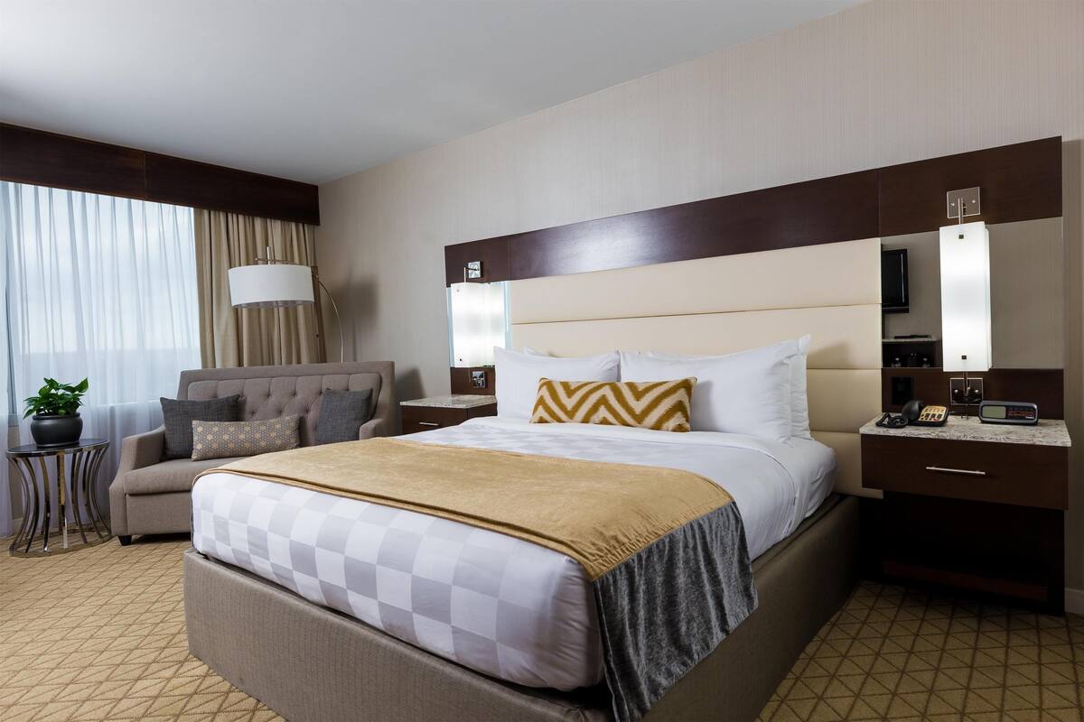 Quarto de hotel com uma cama grande, um sofá, uma mesinha com abajur, uma televisão e um minibar.