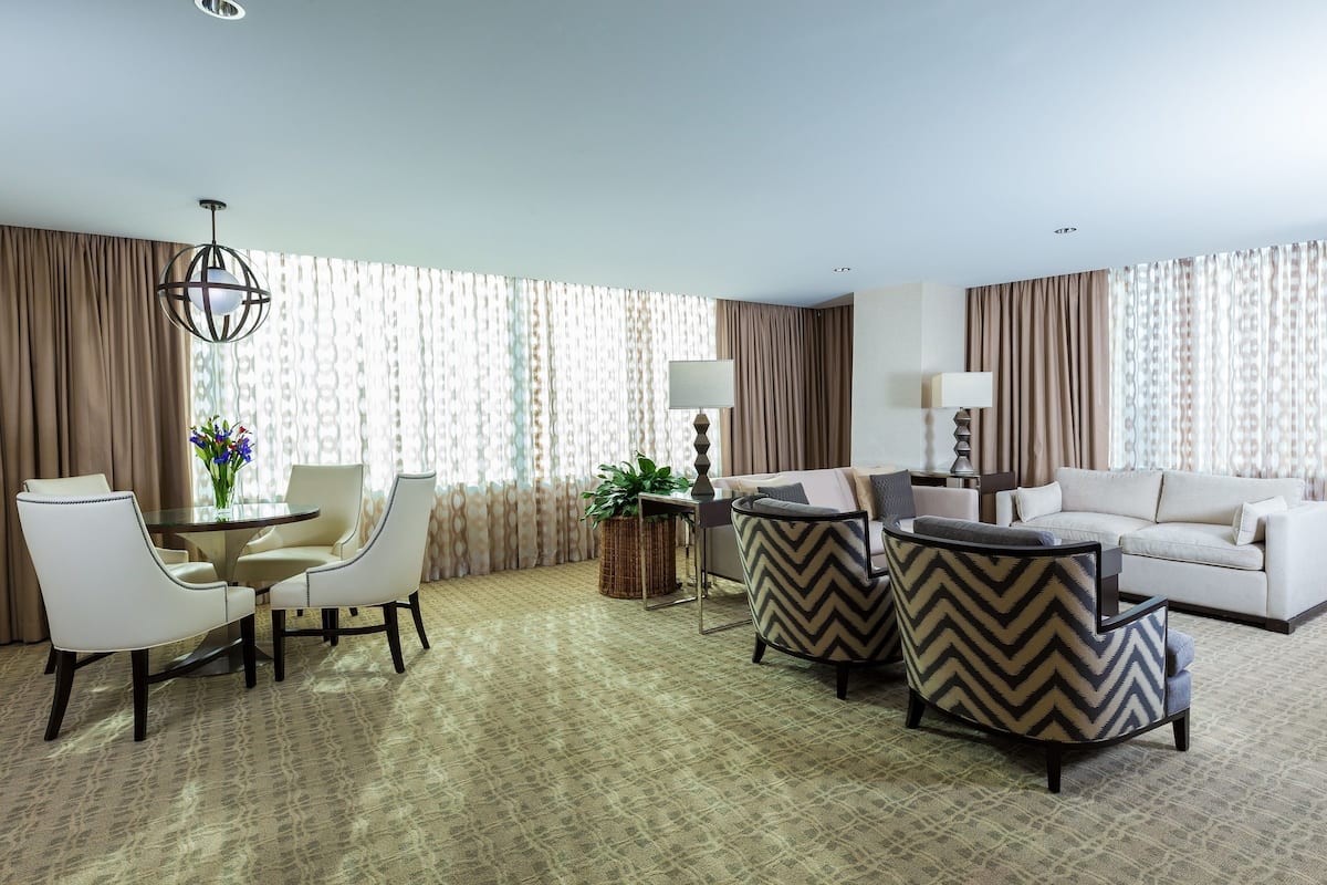 Quarto de hotel com área de refeições, um sofá, uma poltrona estampada, uma luminária e vista para o exterior através de cortinas.