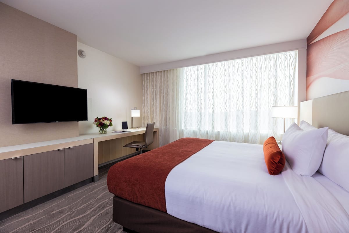 Quarto de hotel com cama, escrivaninha, televisão e janela com cortinas.