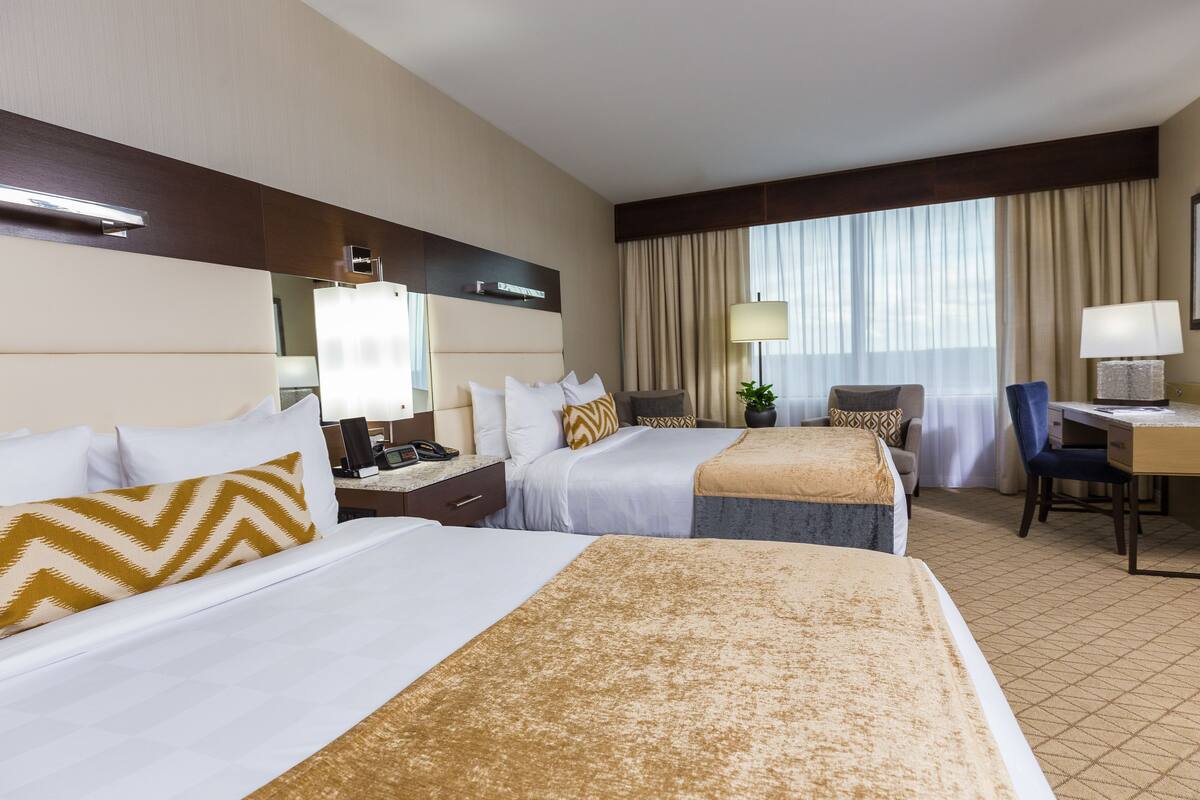 Quarto de hotel com duas camas, uma escrivaninha, uma cadeira, um abajur e uma janela grande com cortinas.