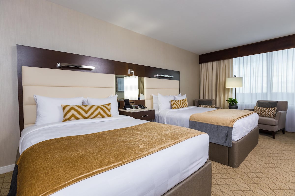 Quarto de hotel com duas camas, uma escrivaninha, uma cadeira e uma janela grande com cortinas.