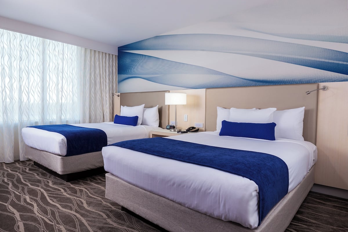 Quarto de hotel com duas camas, uma janela grande com cortinas leves, um mural na parede, uma mesinha de cabeceira com abajur e um tapete estampado.
