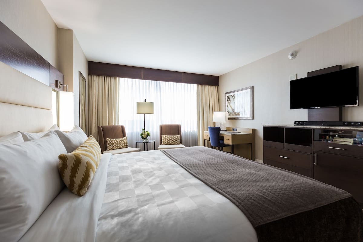 Quarto de hotel com uma cama grande, duas cadeiras, uma escrivaninha, uma televisão e uma cômoda.