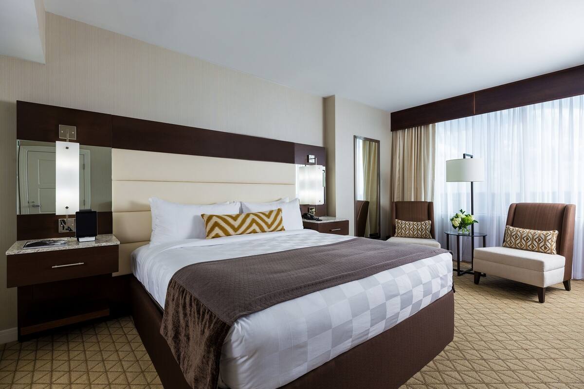 Quarto de hotel com uma cama grande, uma escrivaninha, uma cadeira, um abajur e uma janela com cortinas.