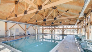 An indoor swimming pool with a wooden ceiling and tiled floor, surrounded by green chairs and tables.