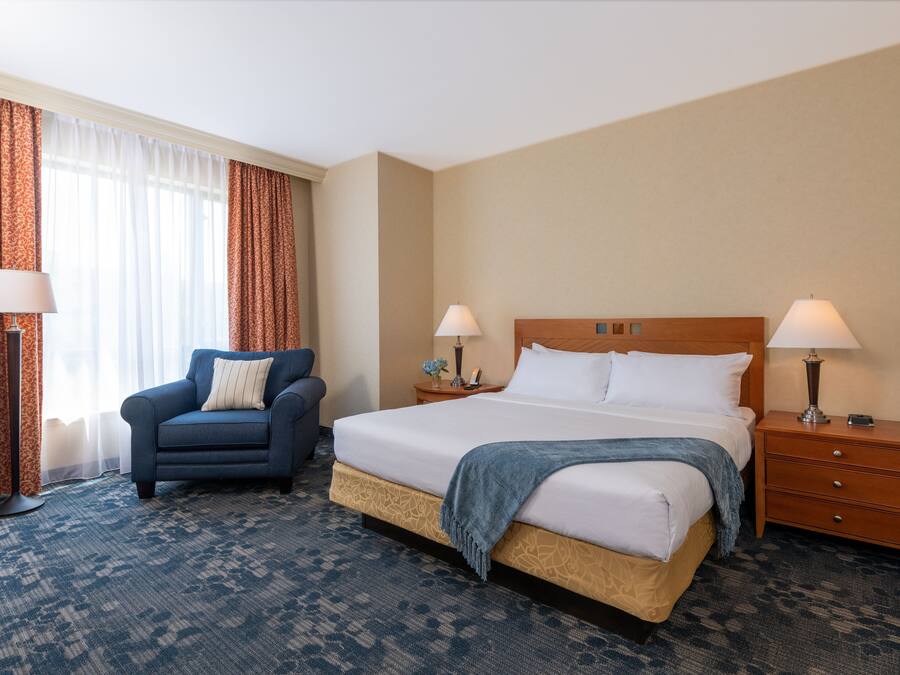 Habitación de hotel con cama, dos mesitas de noche con lámparas, una silla y un ventanal con cortinas.