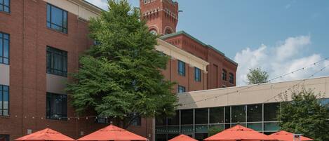 An outdoor patio area with tables and chairs, surrounded by brick buildings and a large tree.