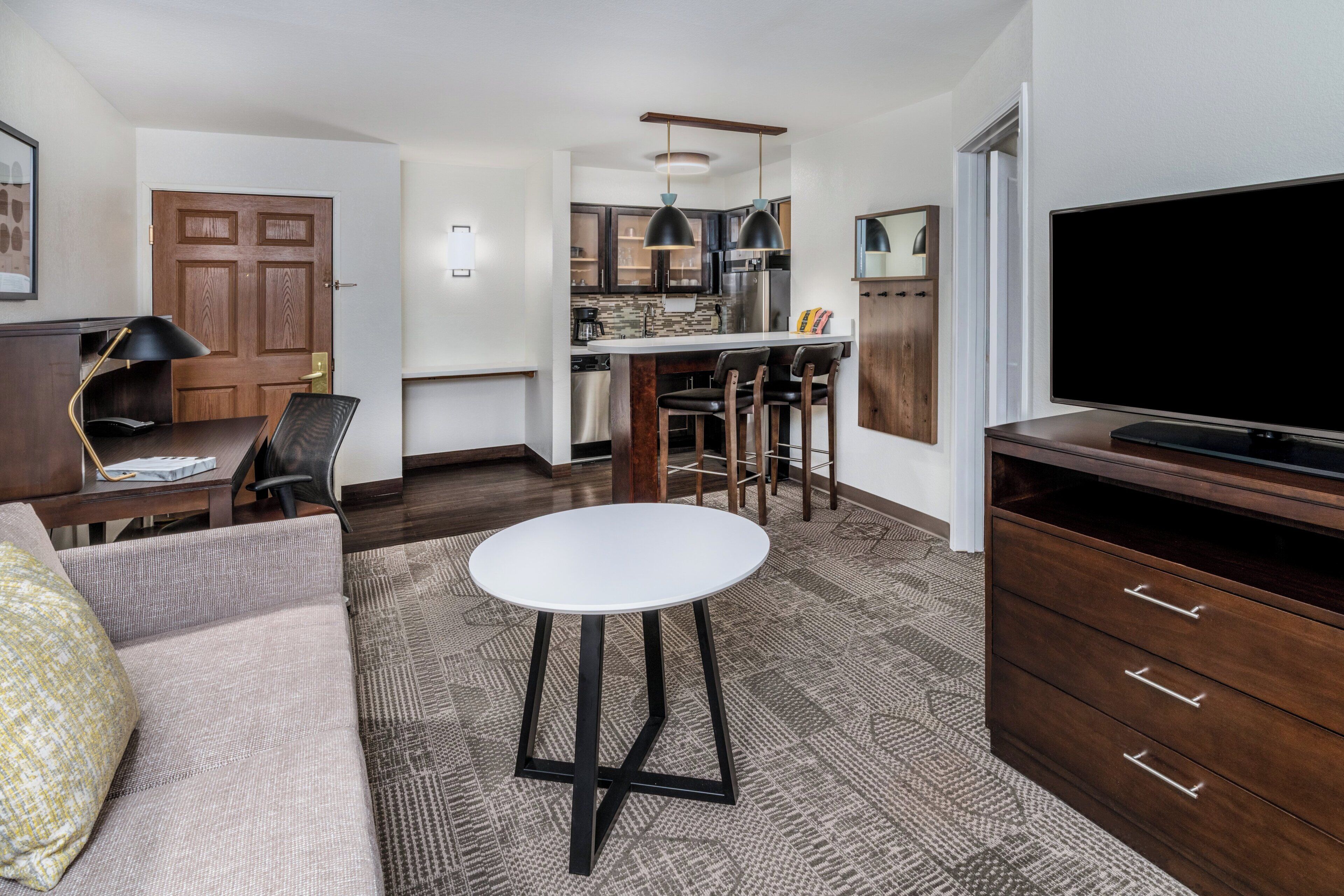 Un salon moderne comprenant un canapé, une table ronde, un téléviseur, un coin cuisine avec des tabourets de bar et un meuble en bois.