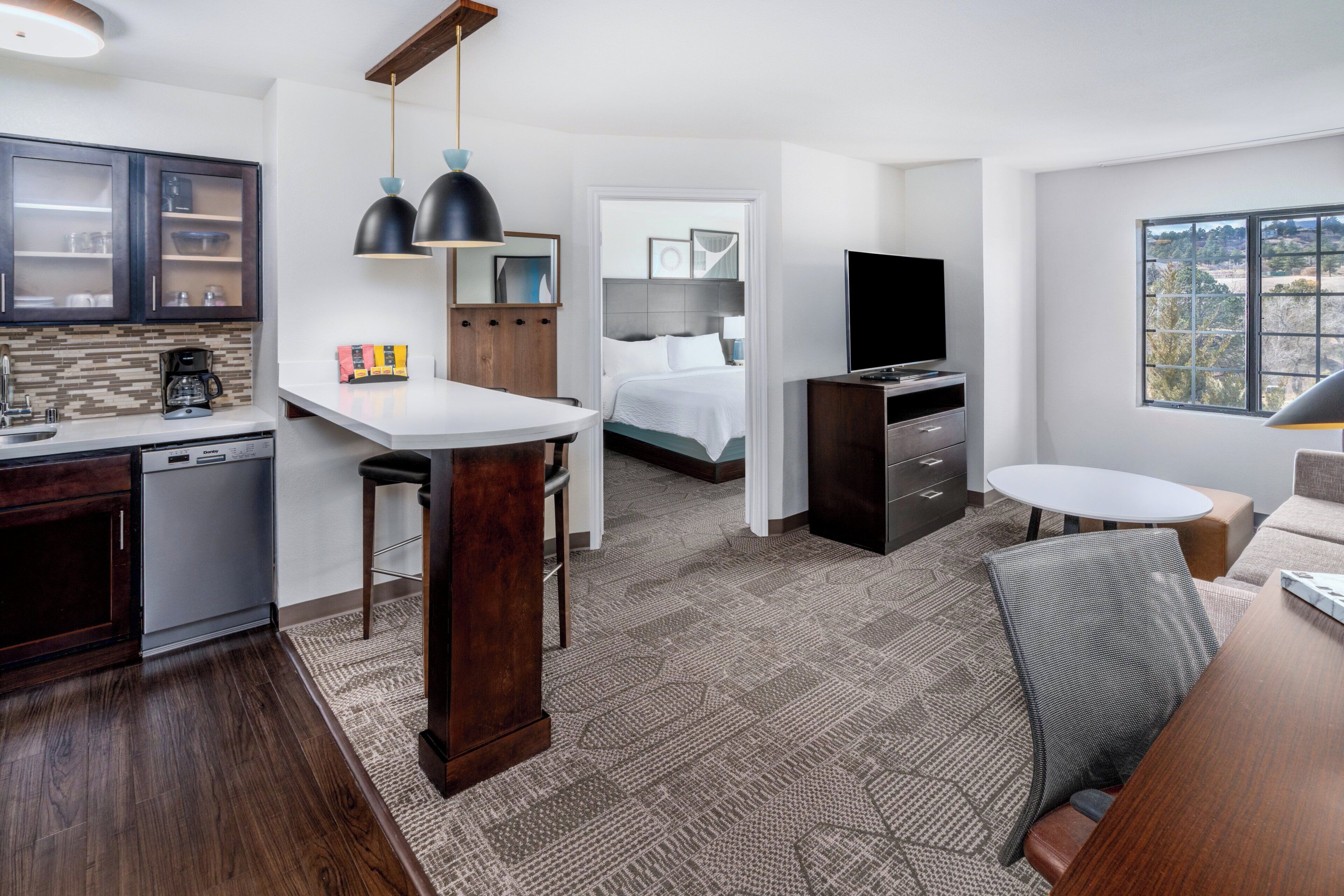 A modern hotel room with a kitchen area, a bed with a view, a TV, and a dining area.