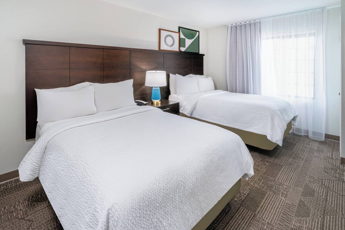 Habitación de hotel con dos camas, una mesita de noche con lámpara, un cuadro enmarcado y una ventana con cortinas.