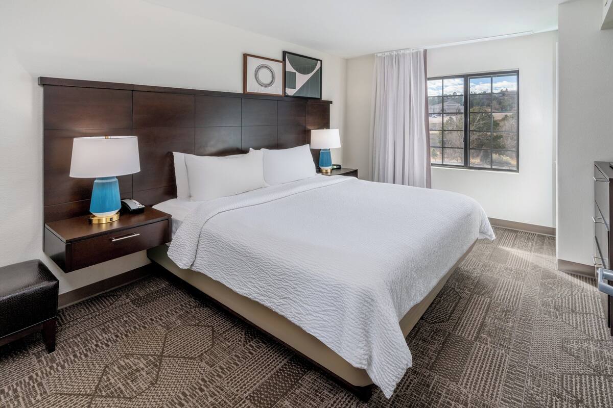 Habitación de hotel con una cama grande, dos mesitas de noche con lámparas, cabecera, ventana con cortinas y vistas al exterior.
