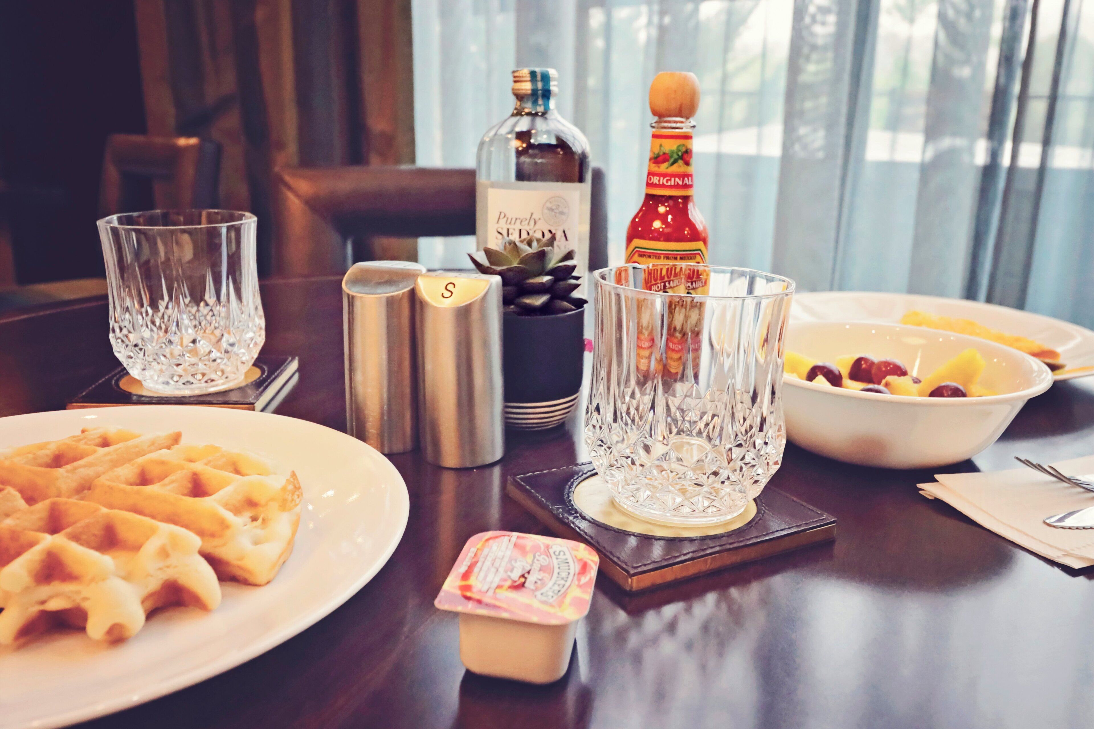 Un desayuno con waffles, un bol de fruta, una botella de sirope y una botella de licor sobre una mesa de madera.