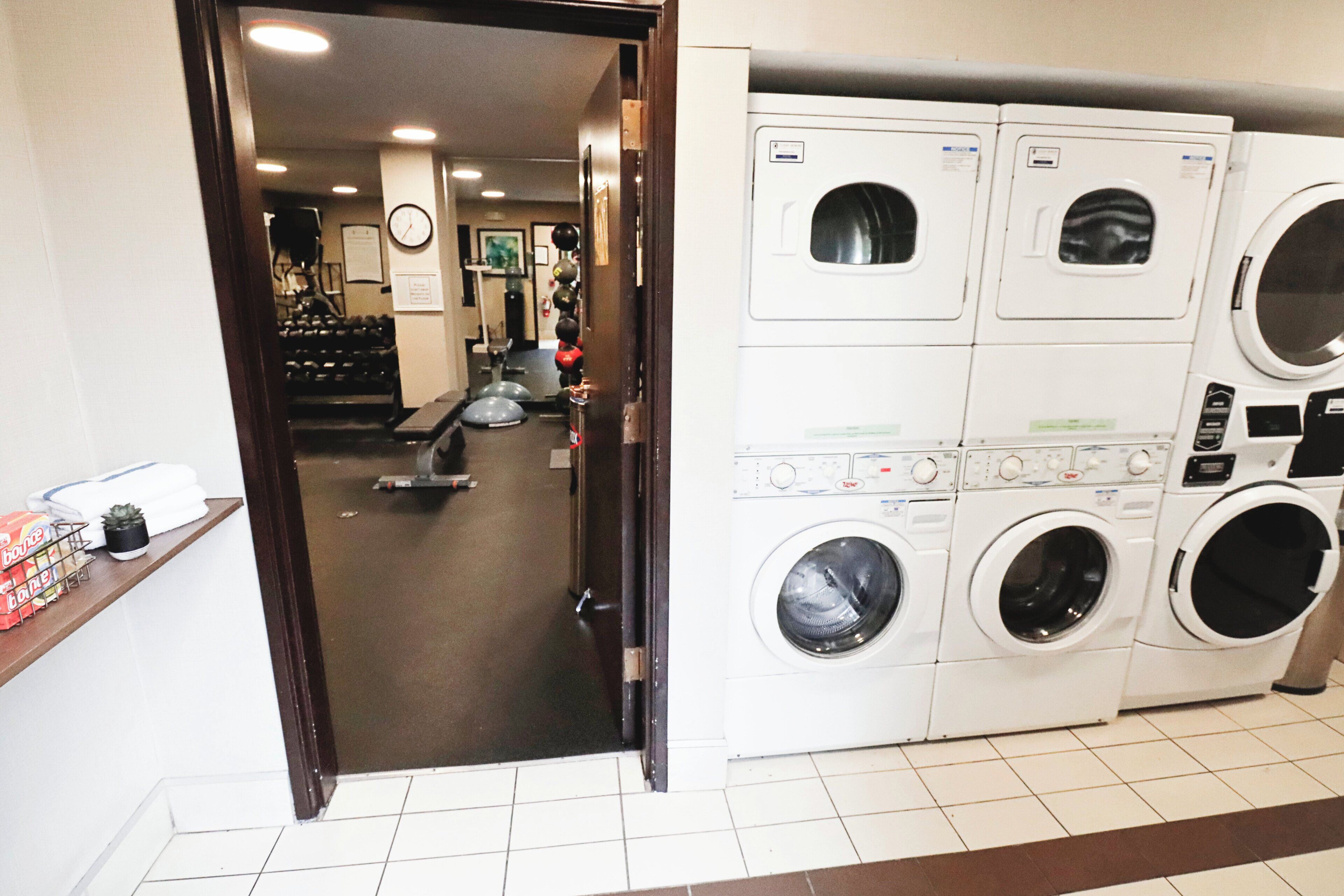 A laundry facility with multiple washing machines and dryers, a clock on the wall, and exercise equipment in the background.