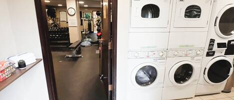 A laundry facility with multiple washing machines and dryers, a clock on the wall, and exercise equipment in the background.