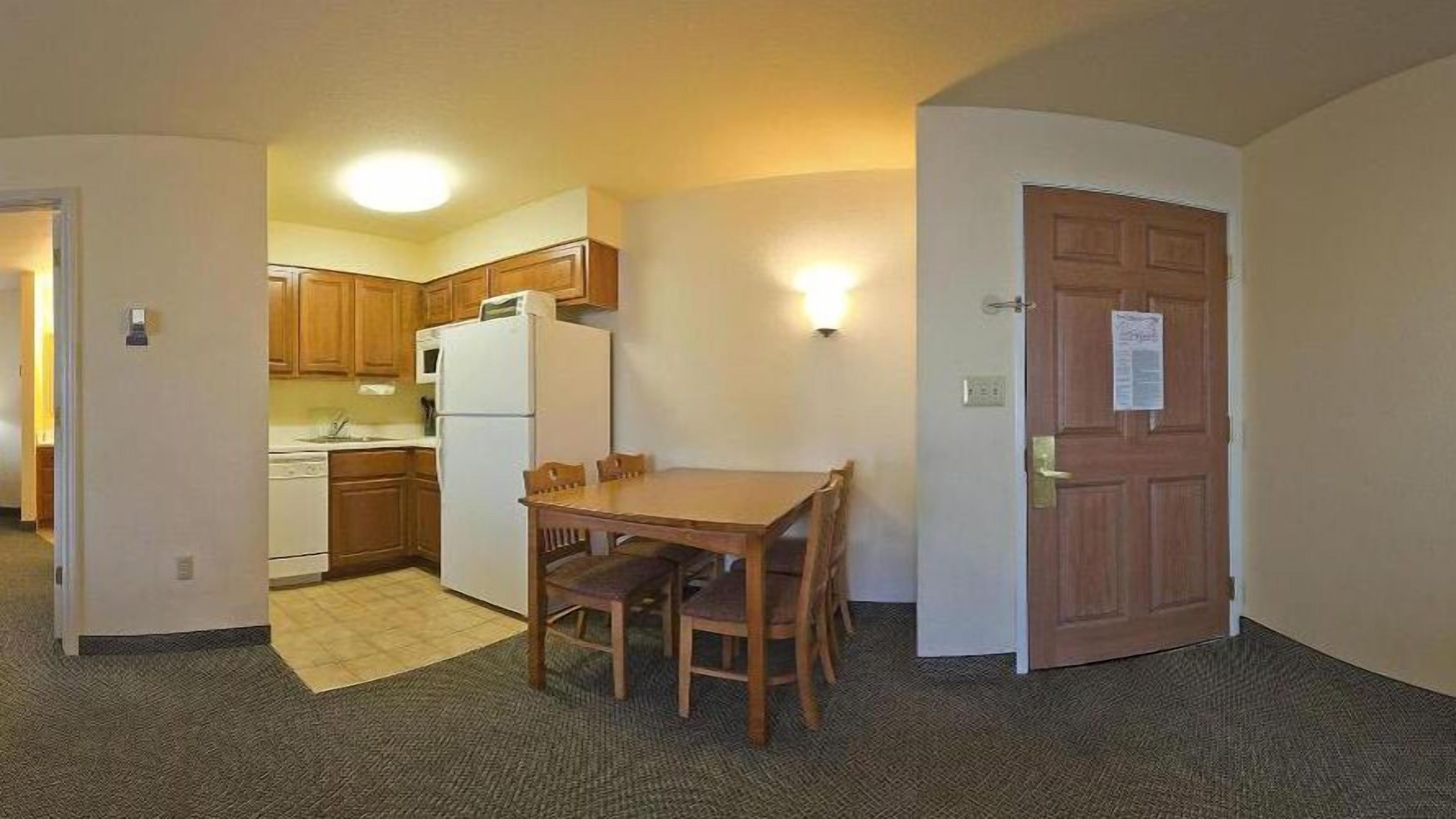 A compact kitchen with wooden cabinets, a refrigerator, and a microwave. There is a dining table with four chairs and a rug on the floor.