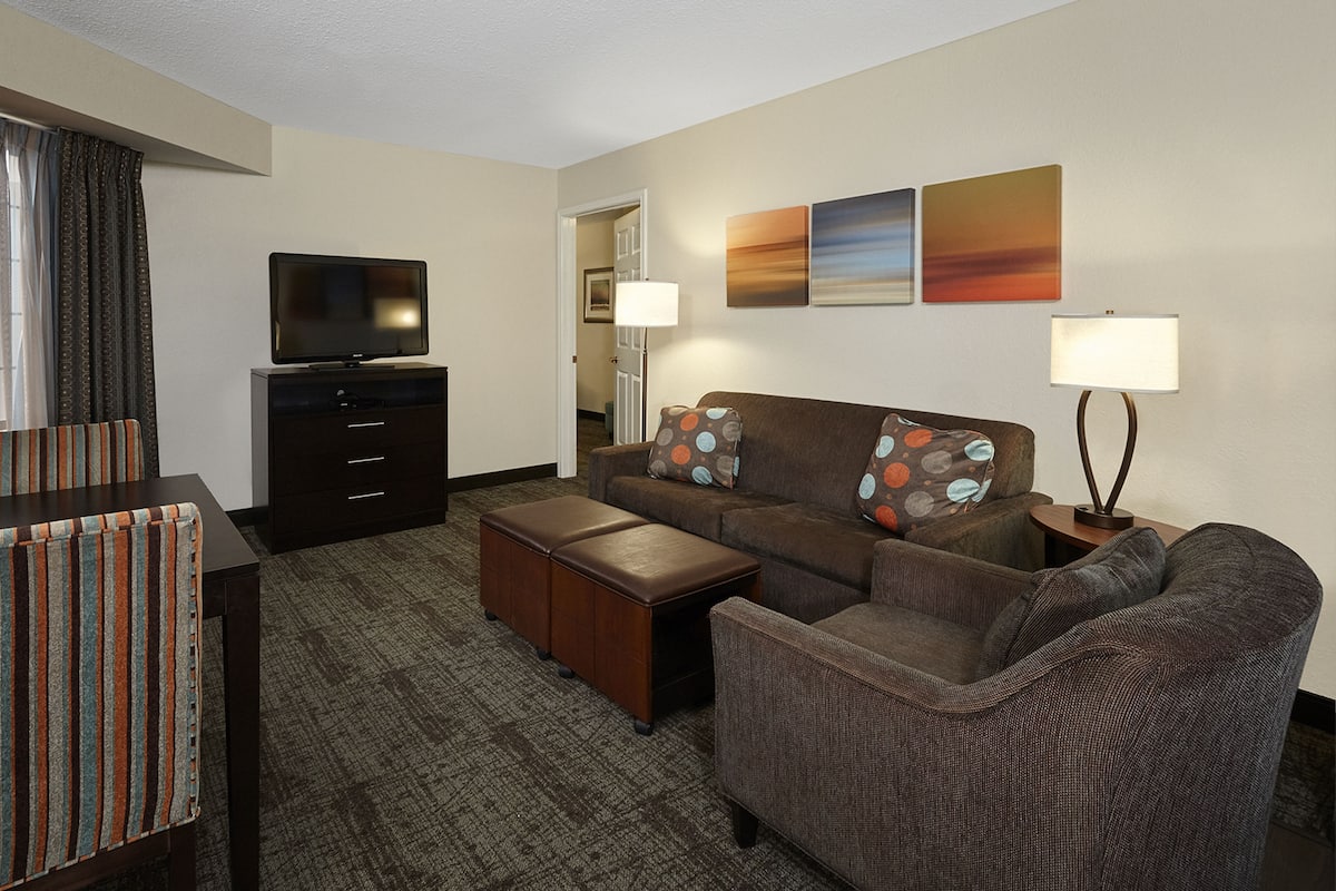A hotel room with a sofa, armchair, ottoman, TV, and artwork on the wall.