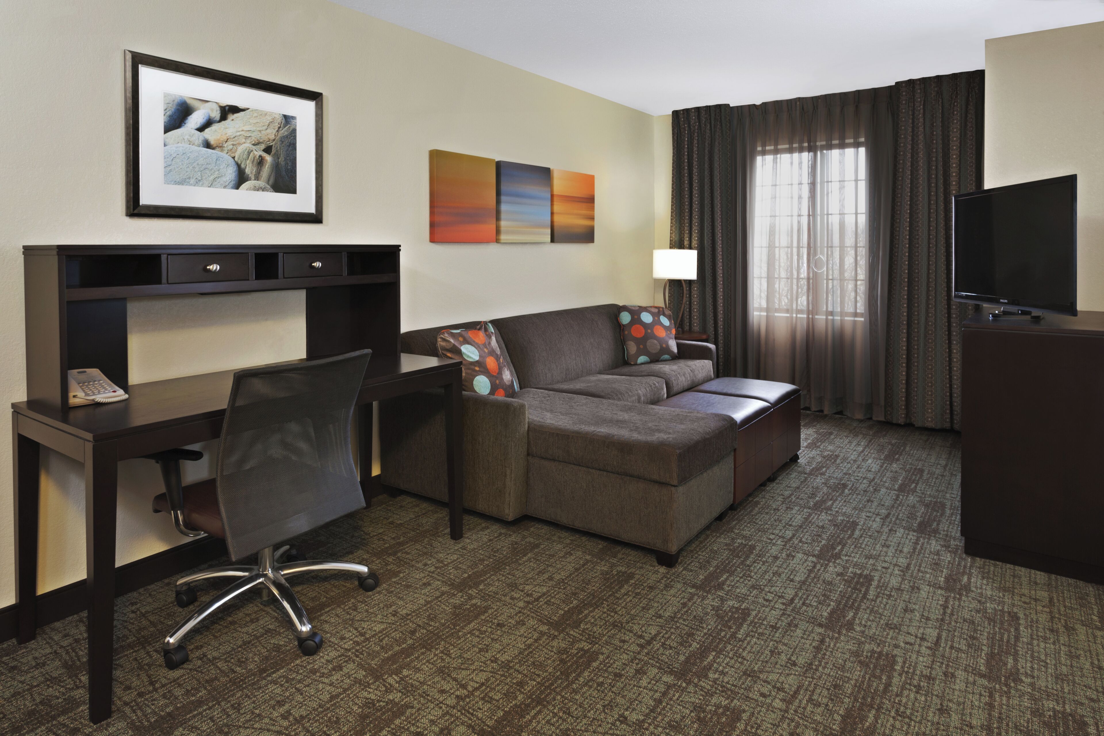 A hotel room with a desk, a sofa, a TV, a lamp, and a window with curtains.