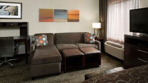 Ein modernes Hotelzimmer mit Ecksofa, Flachbildfernseher, Schreibtisch und großem Fenster mit Vorhängen.