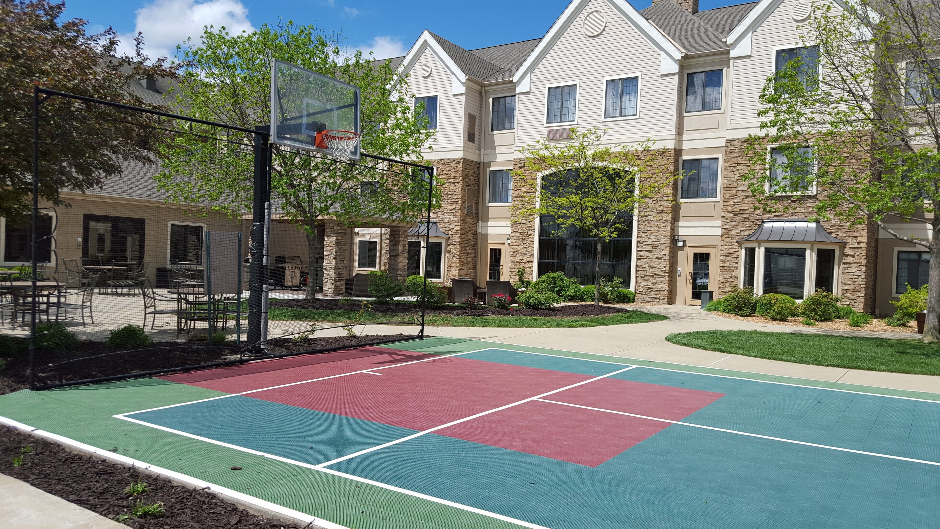An outdoor basketball court with a net, surrounded by a building with stone and wood siding, and a patio area with outdoor seating.