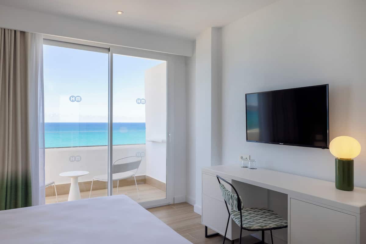 A hotel room with a bed, a desk, a chair, a TV, and a balcony with a view of the sea.