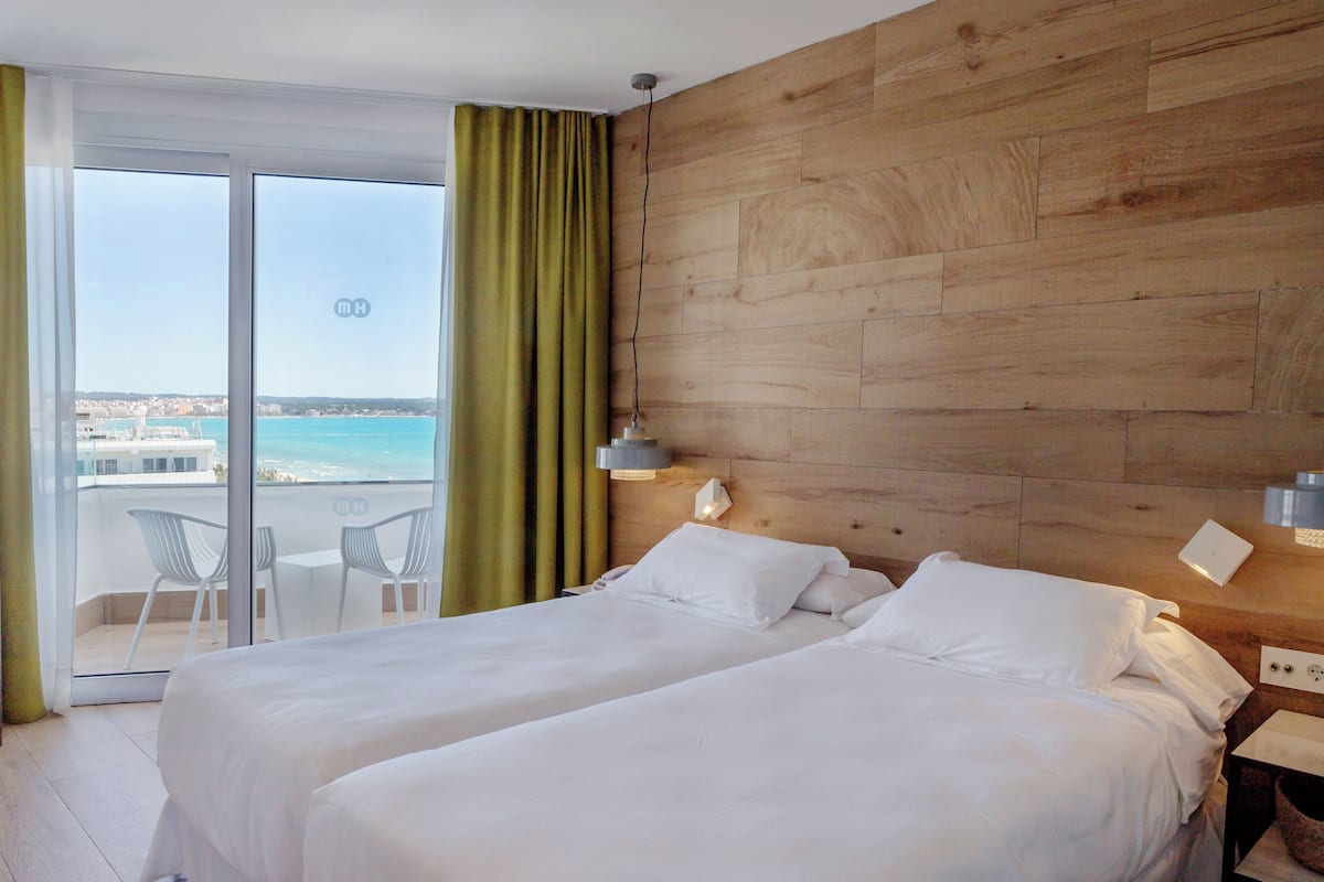 A hotel room with a wooden headboard, two beds, a balcony with a view of the sea, and a table with chairs.