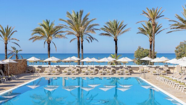 Zona de piscina con sillas de descanso y sombrillas, palmeras y vistas al mar.