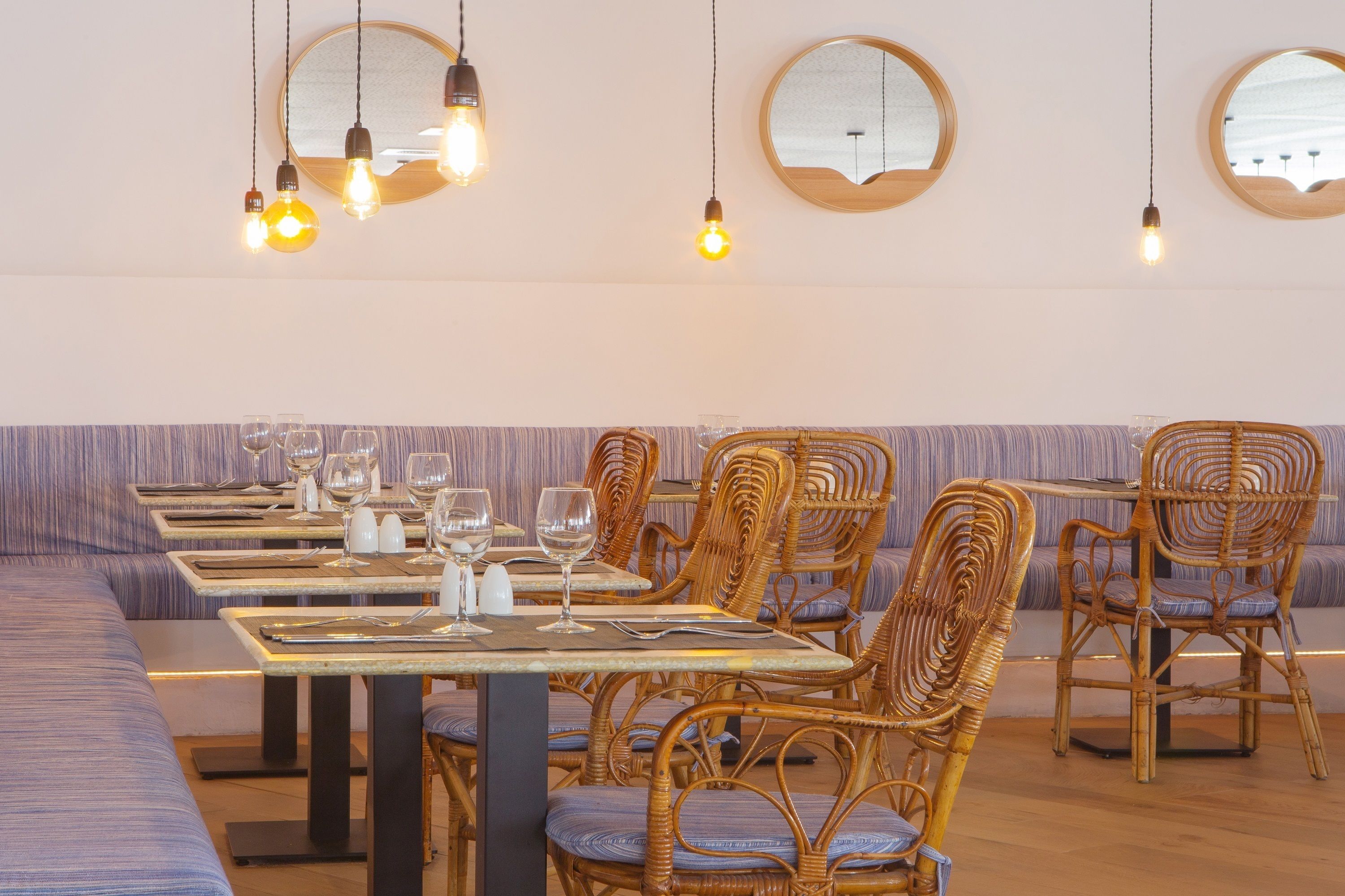 A restaurant with wicker chairs and tables, hanging light fixtures, and a striped bench.