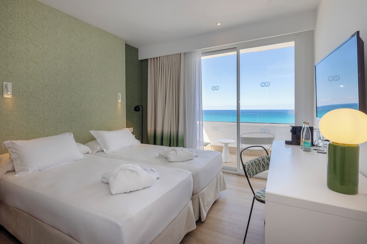 A hotel room with two beds, a desk, a chair, and a view of the ocean.