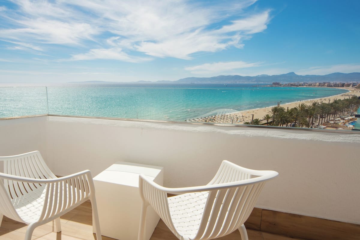 A balcony with two white chairs and a small table, overlooking a beach and the sea.