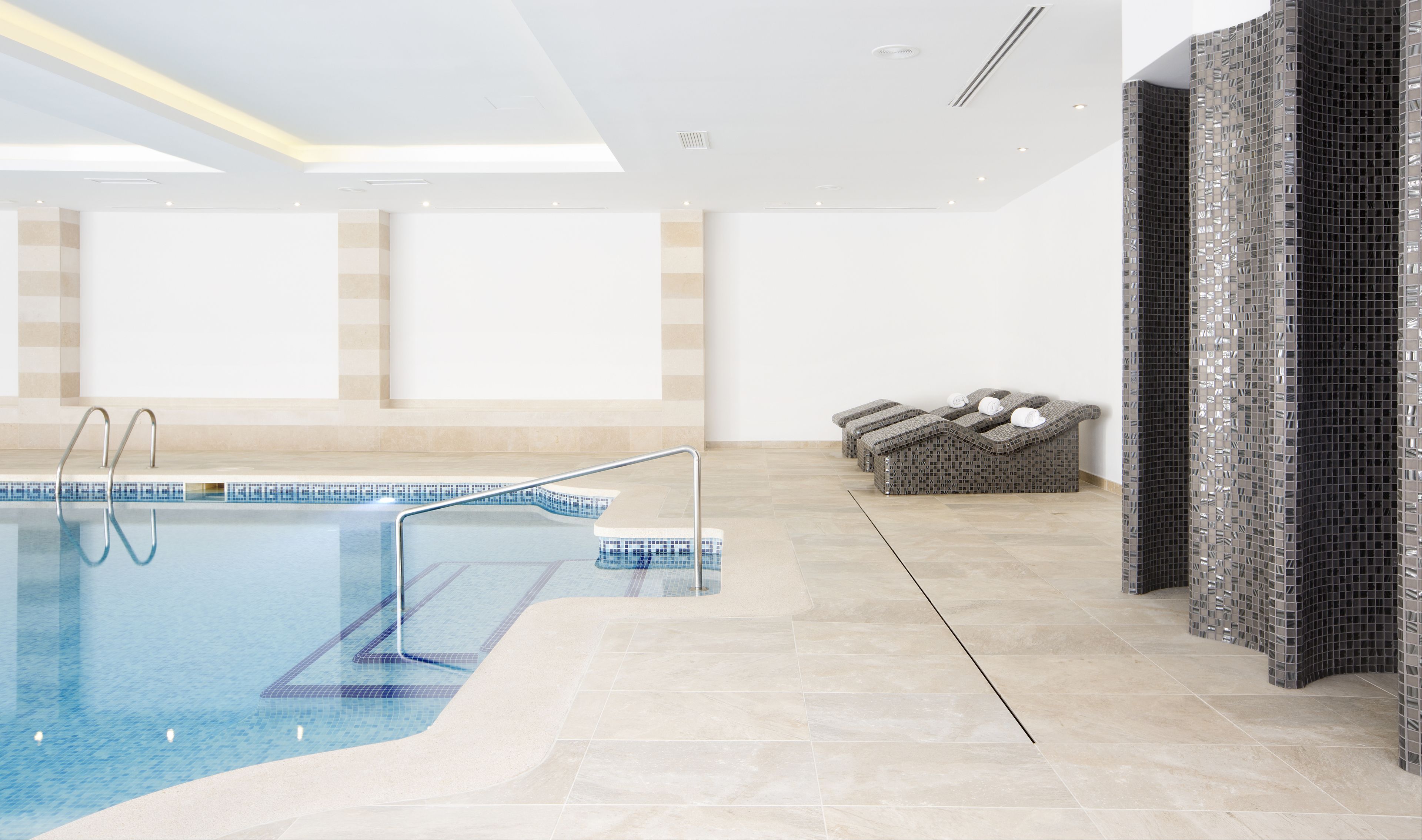 Una piscina interior amb aigua cristal·lina, una escala metàl·lica i un paviment de rajoles. Hi ha seients de relaxació amb tovalloles a una paret amb un disseny de mosaics.