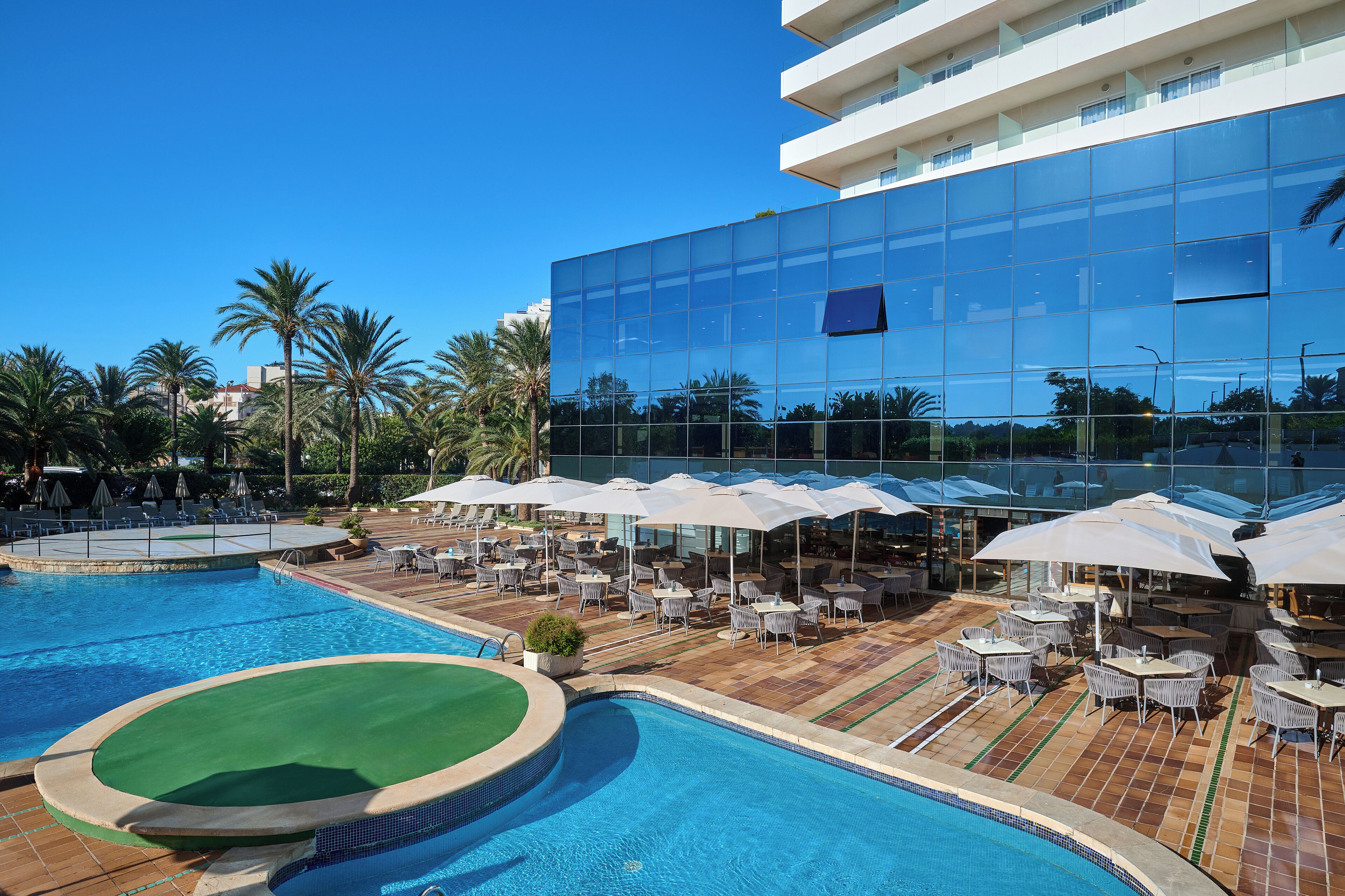 A hotel pool area with a large glass building, outdoor seating, and palm trees.