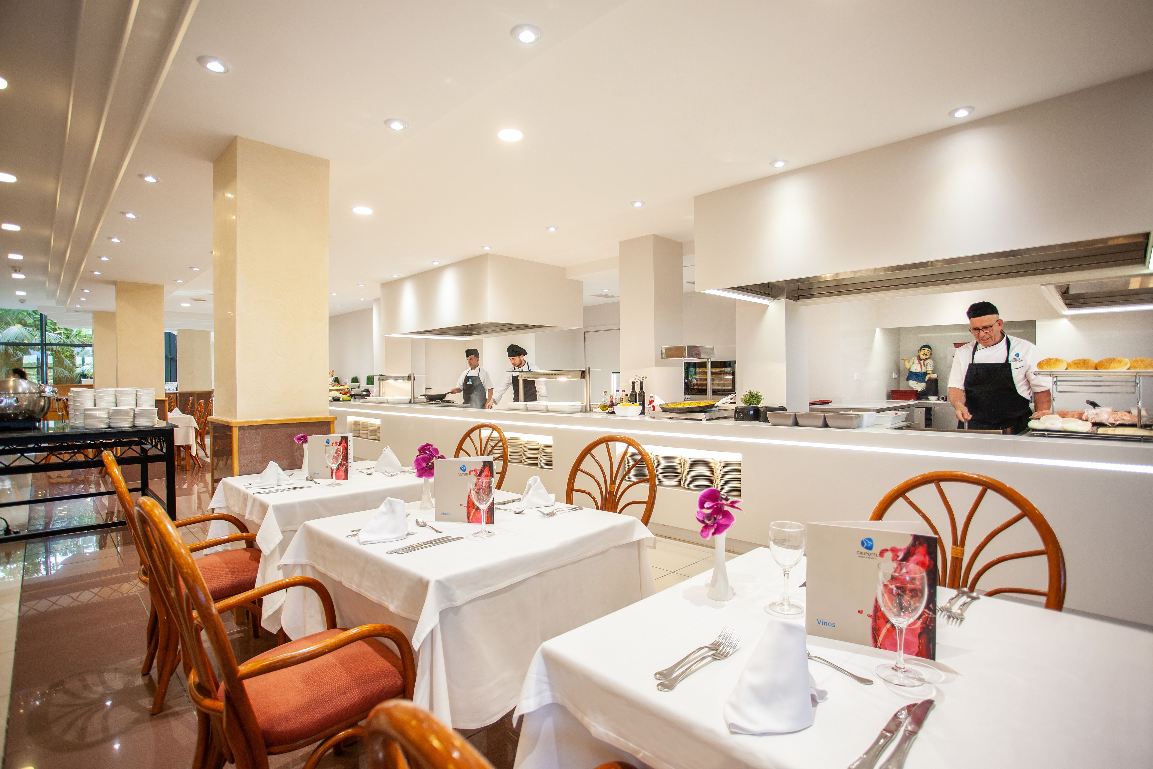 Een modern restaurant met witte tafels en stoelen, een keuken en uitzicht op het buitengebied.
