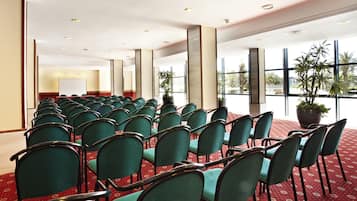 Un salón de conferencias con filas de sillas verdes, ventanas amplias y plantas en macetas.