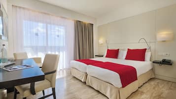 A hotel room with a large bed, a desk with a chair, a bedside table with a lamp, and a window with curtains.