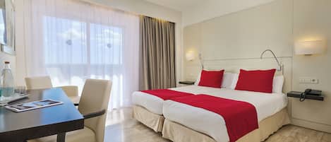 Habitación de hotel con una cama grande, un escritorio con una silla, una mesita de noche con una lámpara y una ventana con cortinas.