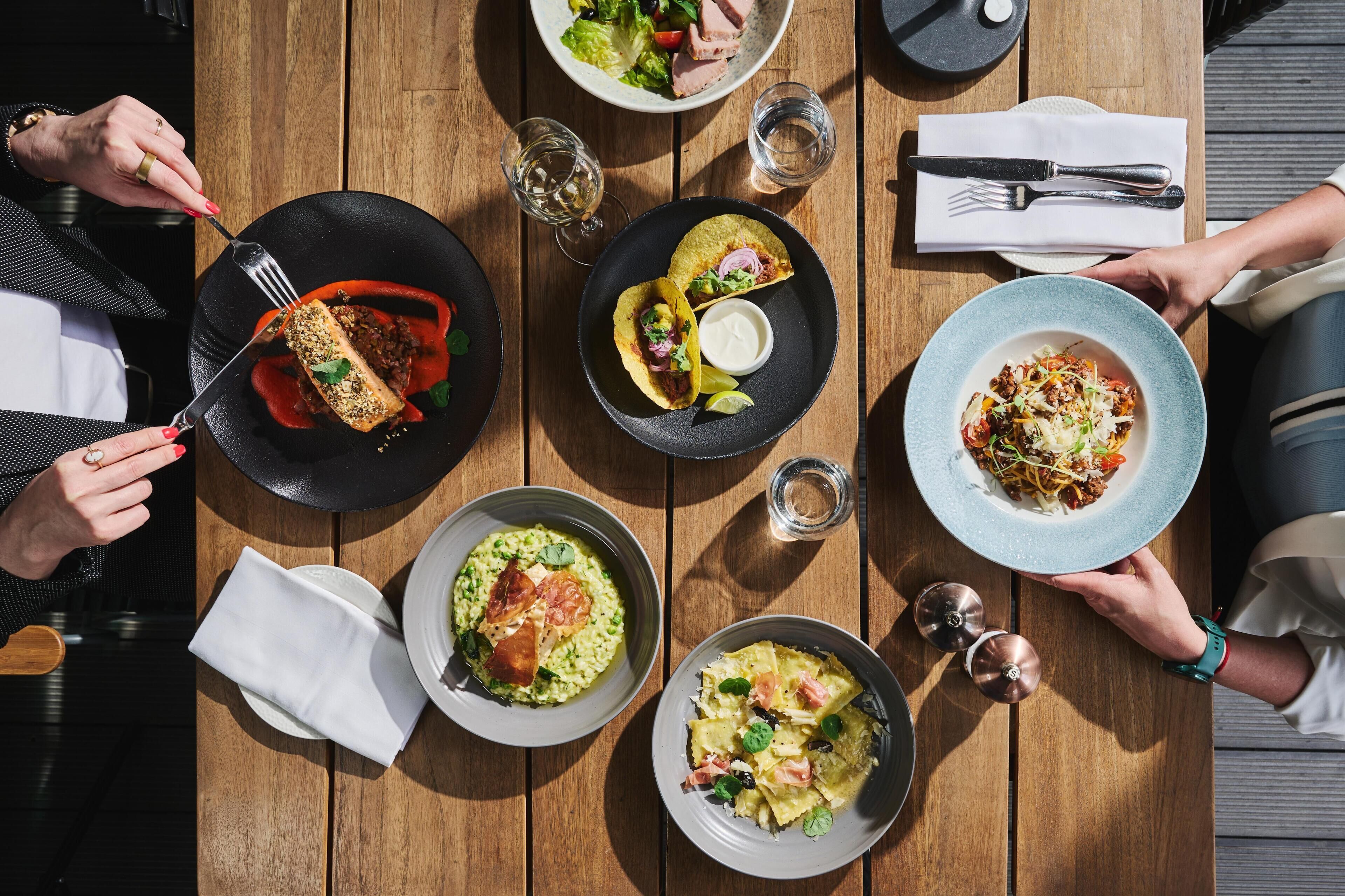 Et træbord med forskellige retter, heriblandt tacos, pasta og en salat, der indikerer et spiseområde.