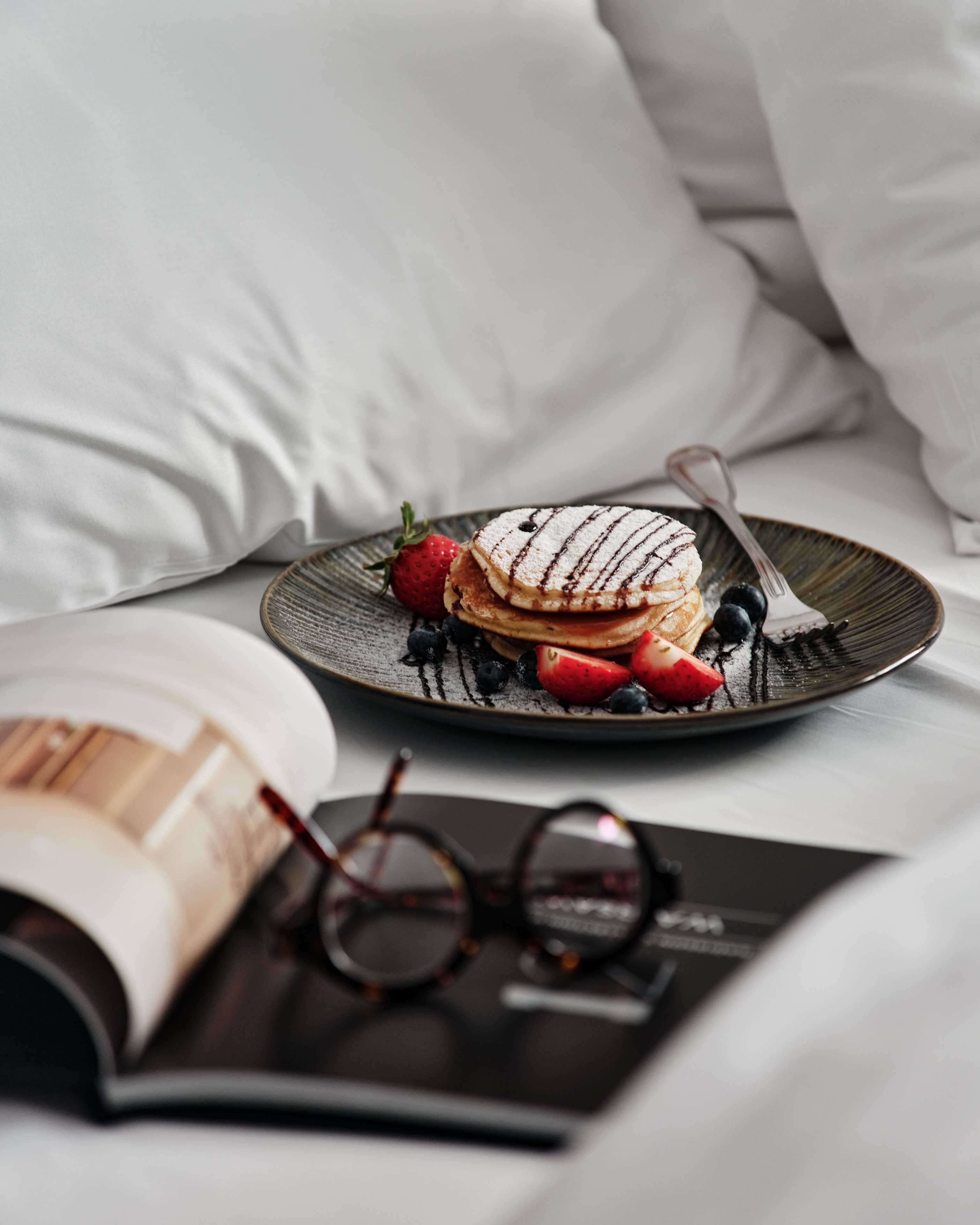 Uma pilha de panquecas com frutas vermelhas e açúcar em pó sobre uma cama com roupa de cama branca e uma revista.