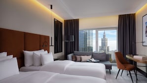 A hotel room with two beds, a sofa, a small table, and a chair. There is a large window with a view of city buildings. - Radisson Collection Hotel, Warsaw (Warsaw)