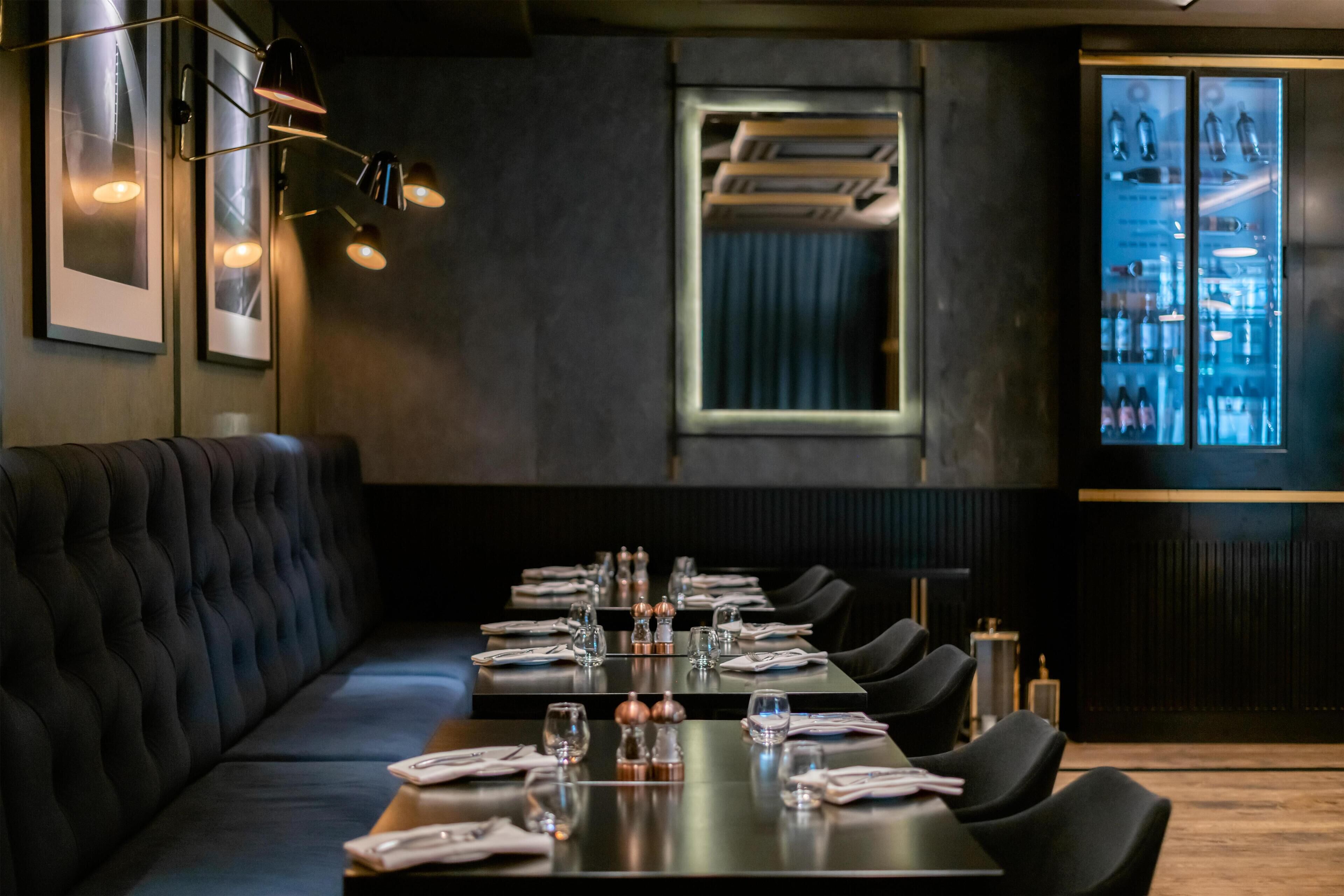 Un restaurante con una larga mesa dispuesta para comer, asientos acolchados, iluminación colgante y un área de bar.