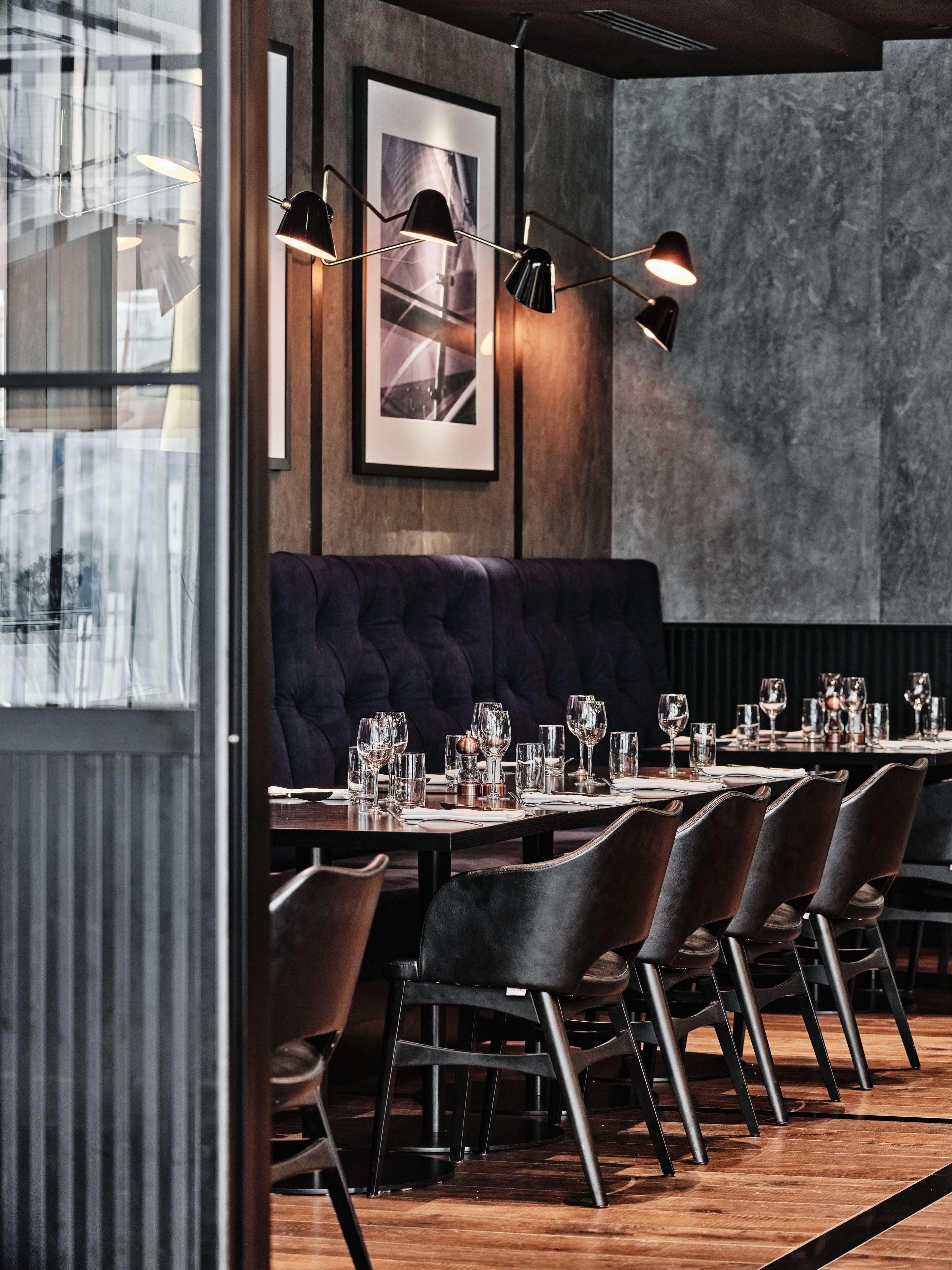 Een restaurant met een donker houten vloer, een ruime gewatteerde zithoek en een lange tafel gedekt met glazen en bestek.