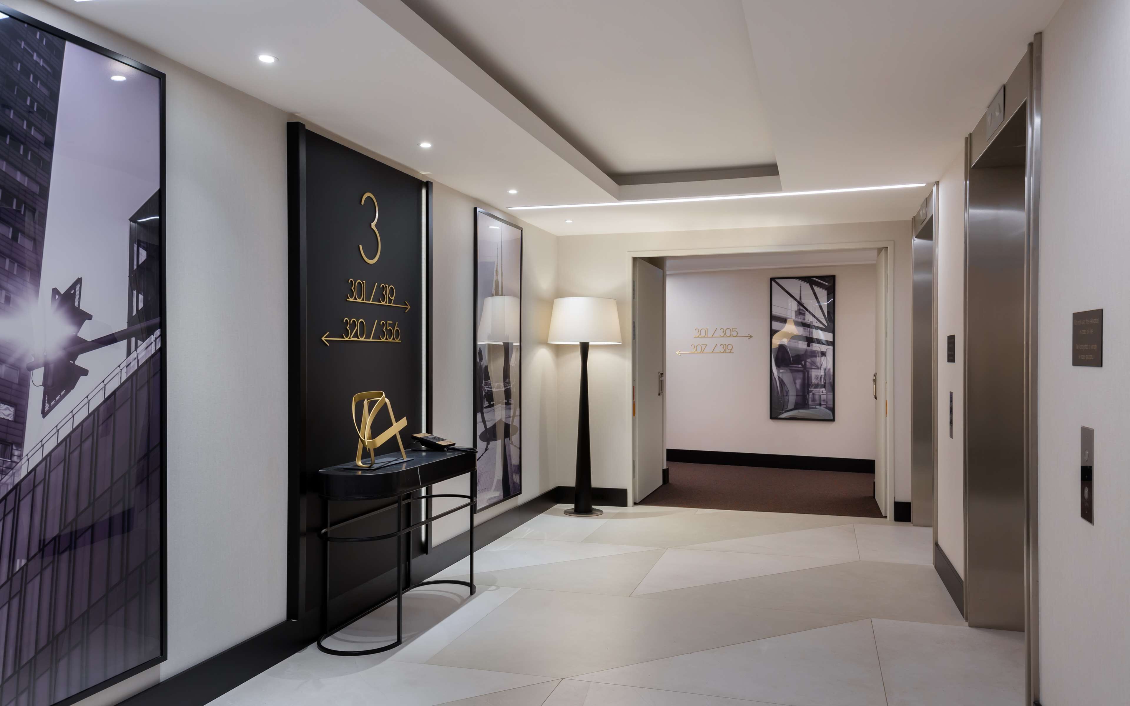 A modern hotel corridor with directional signs, a floor lamp, and a view of the building outside.