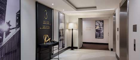 A modern hotel corridor with directional signs, a floor lamp, and a view of the building outside.