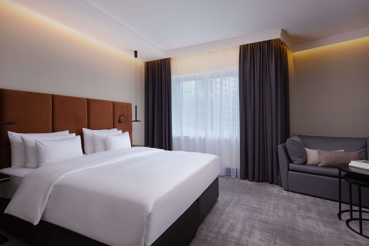 Habitación de hotel con una cama grande, un sofá, una mesita y una ventana con cortinas.