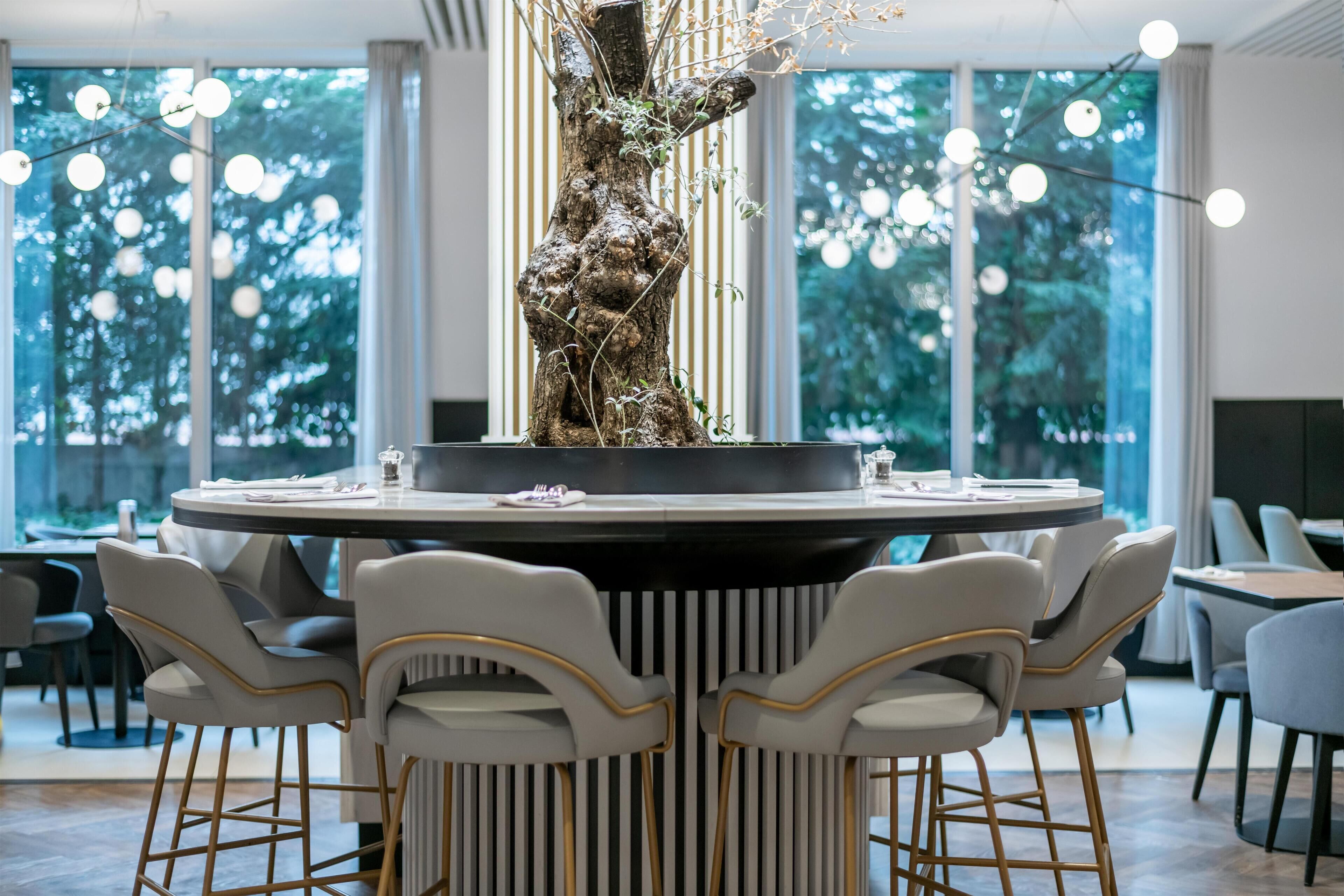 Una mesa redonda para comer con seis sillas, una escultura de árbol en el centro y vistas a árboles a través de amplias ventanas.