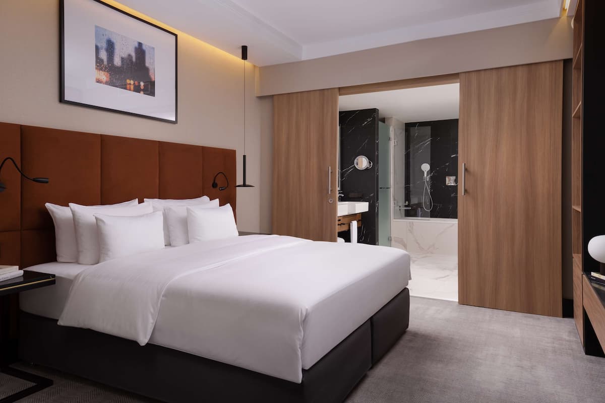 Una habitación de hotel moderna con una cama grande, una mesita de noche con lámpara, un cuadro en la pared y vistas al baño.