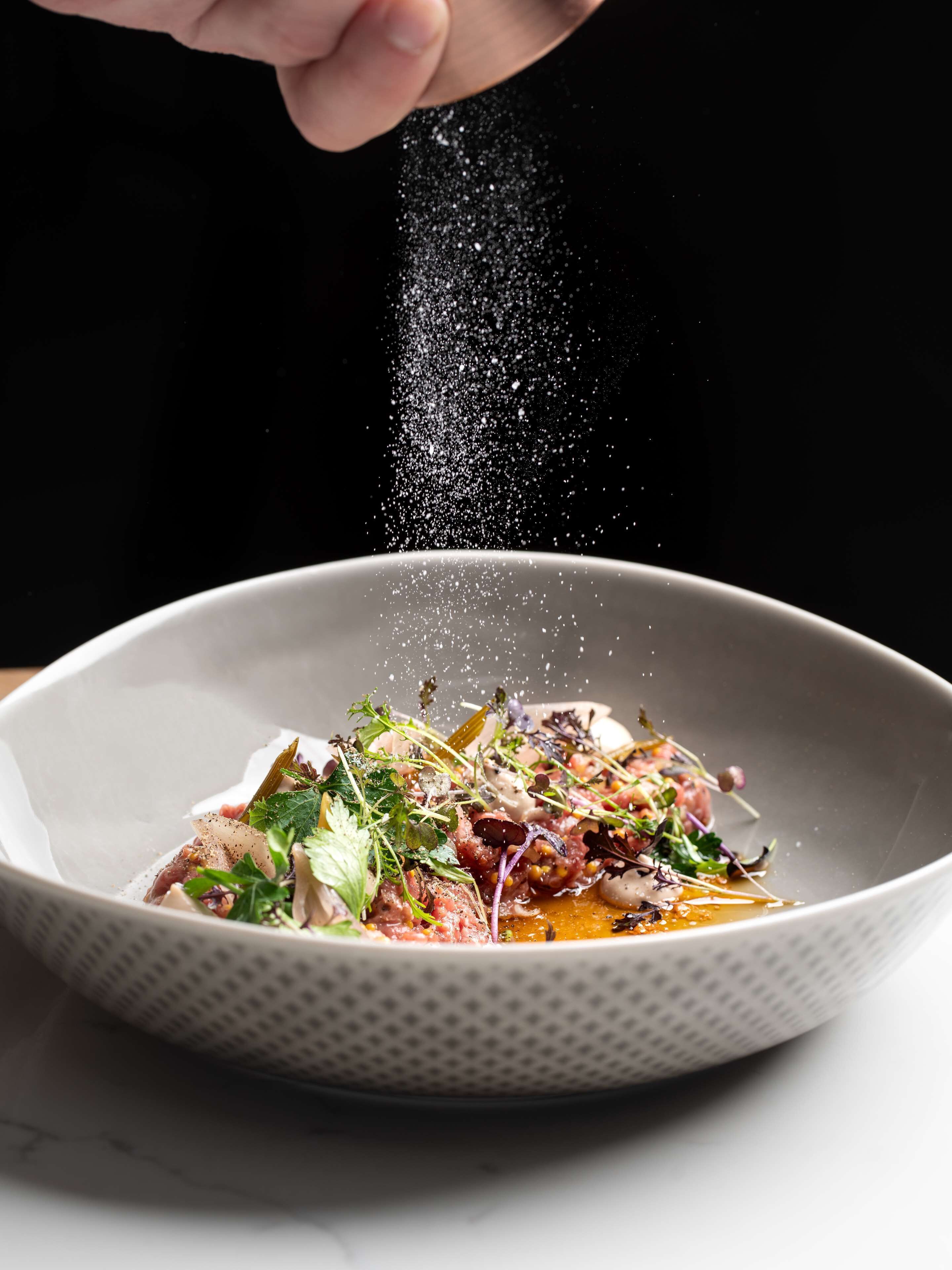 Een schaal eten met diverse ingrediënten, terwijl er kruiden over gestrooid worden.