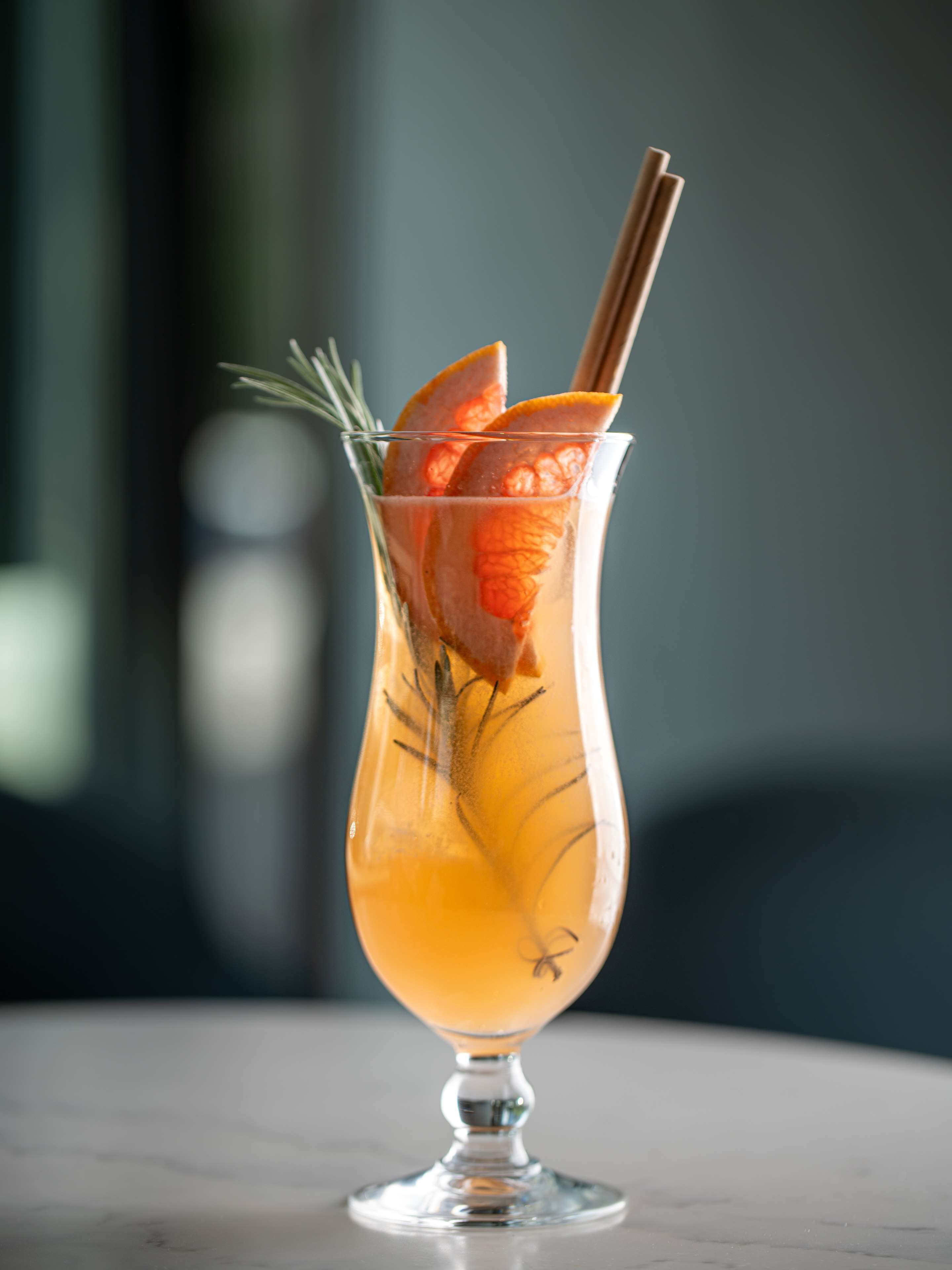 A cocktail glass with a stem, garnished with orange slices and a sprig of rosemary, placed on a marble surface.