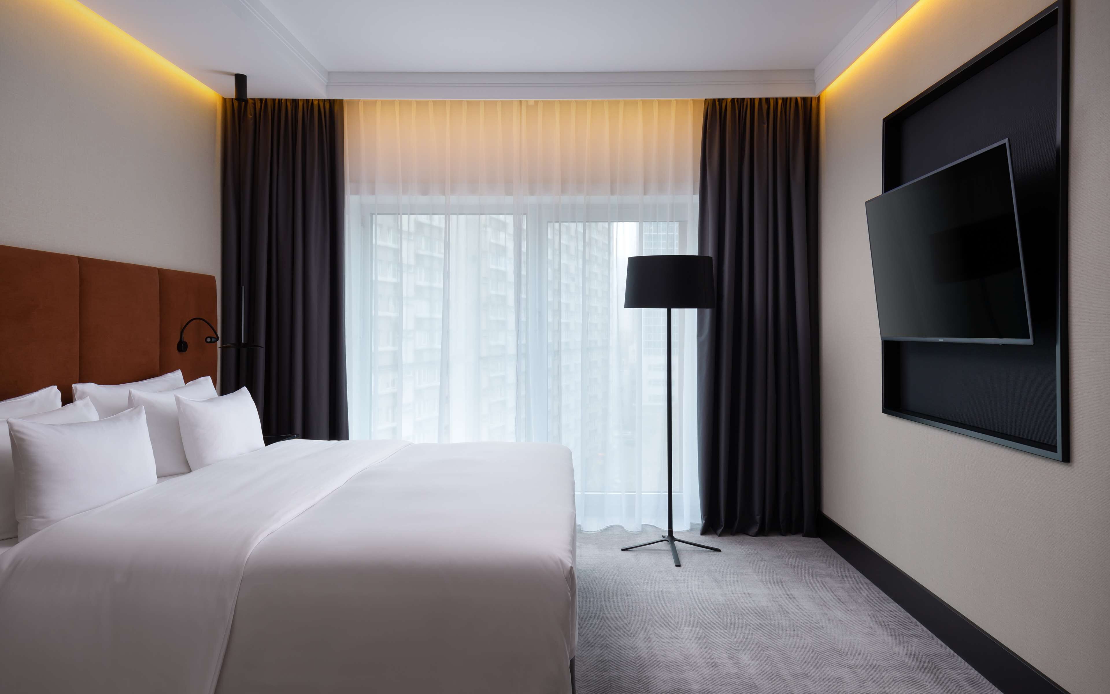 A hotel room with a large bed, a flat-screen TV mounted on the wall, a floor lamp, and a view of the city through the window.