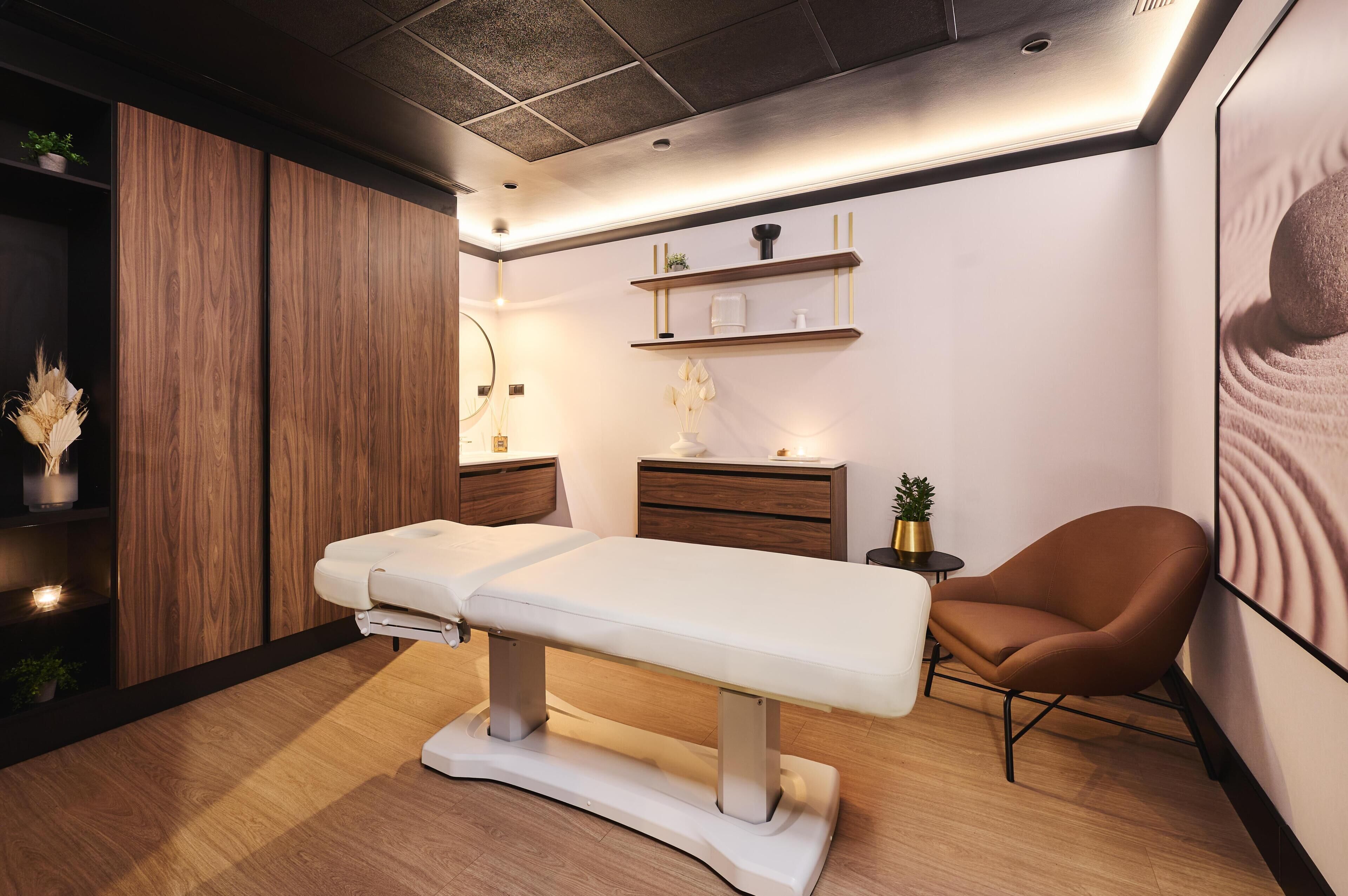 A modern examination room with a wooden panel wall, a white examination table, a brown chair, and a small shelf with decorative items.